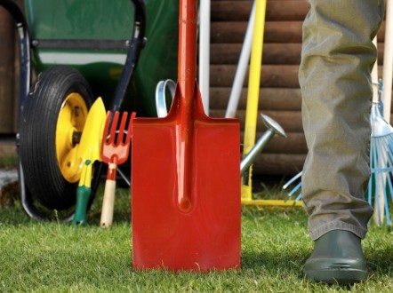 A man is beige trousers and green wellies standing next to a red spade and green wheelbarrow