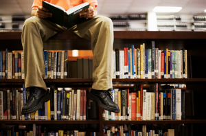 Man sitting on top of library book shelf reading a book.